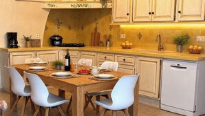 Una cucina con tavolo in legno, sedie bianche, lavastoviglie e un piano di lavoro con diversi oggetti.