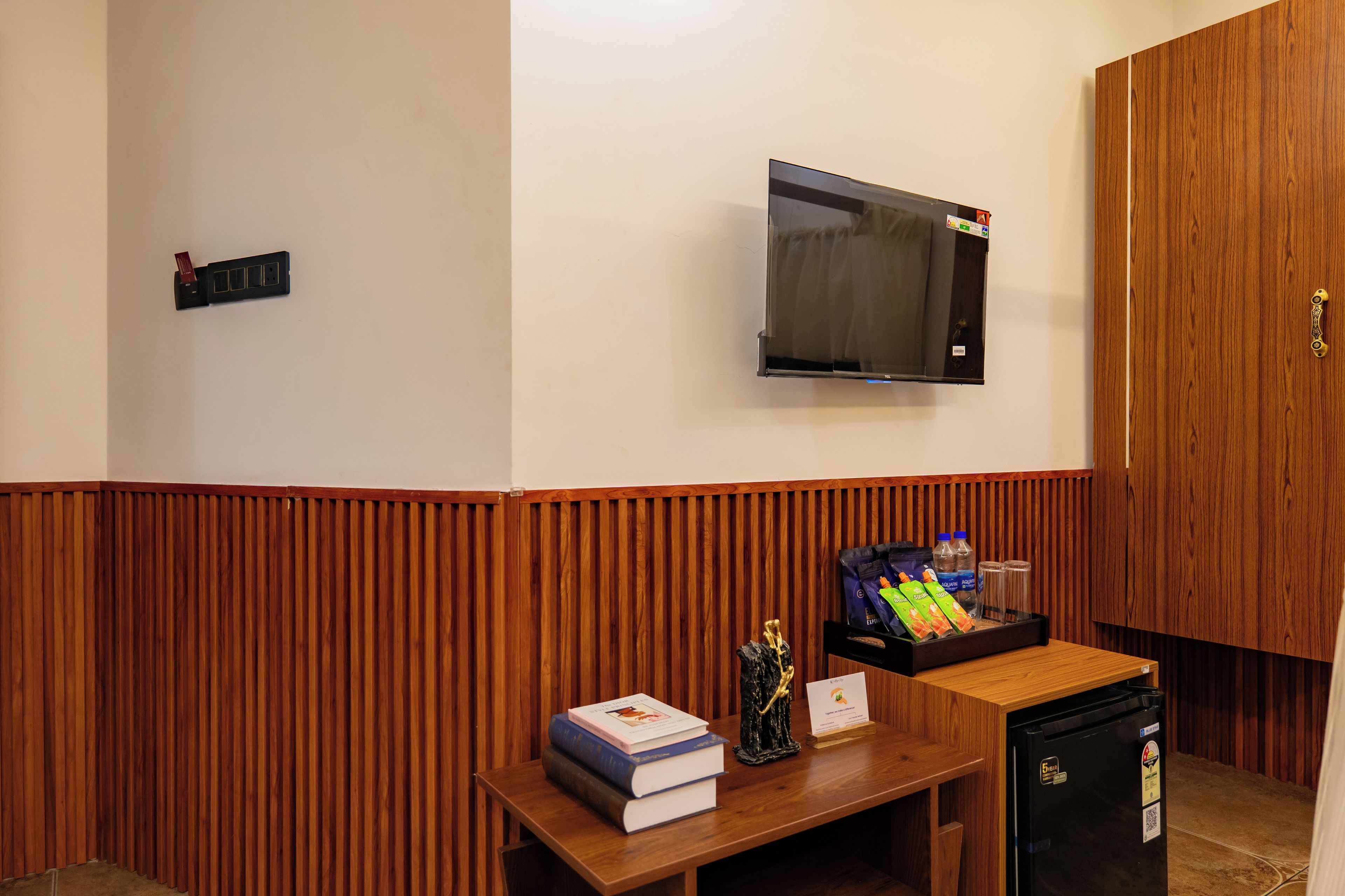 Una habitación con pared de paneles de madera, un televisor de pantalla plana, una mesita con libros y un adorno, y un refrigerador pequeño.