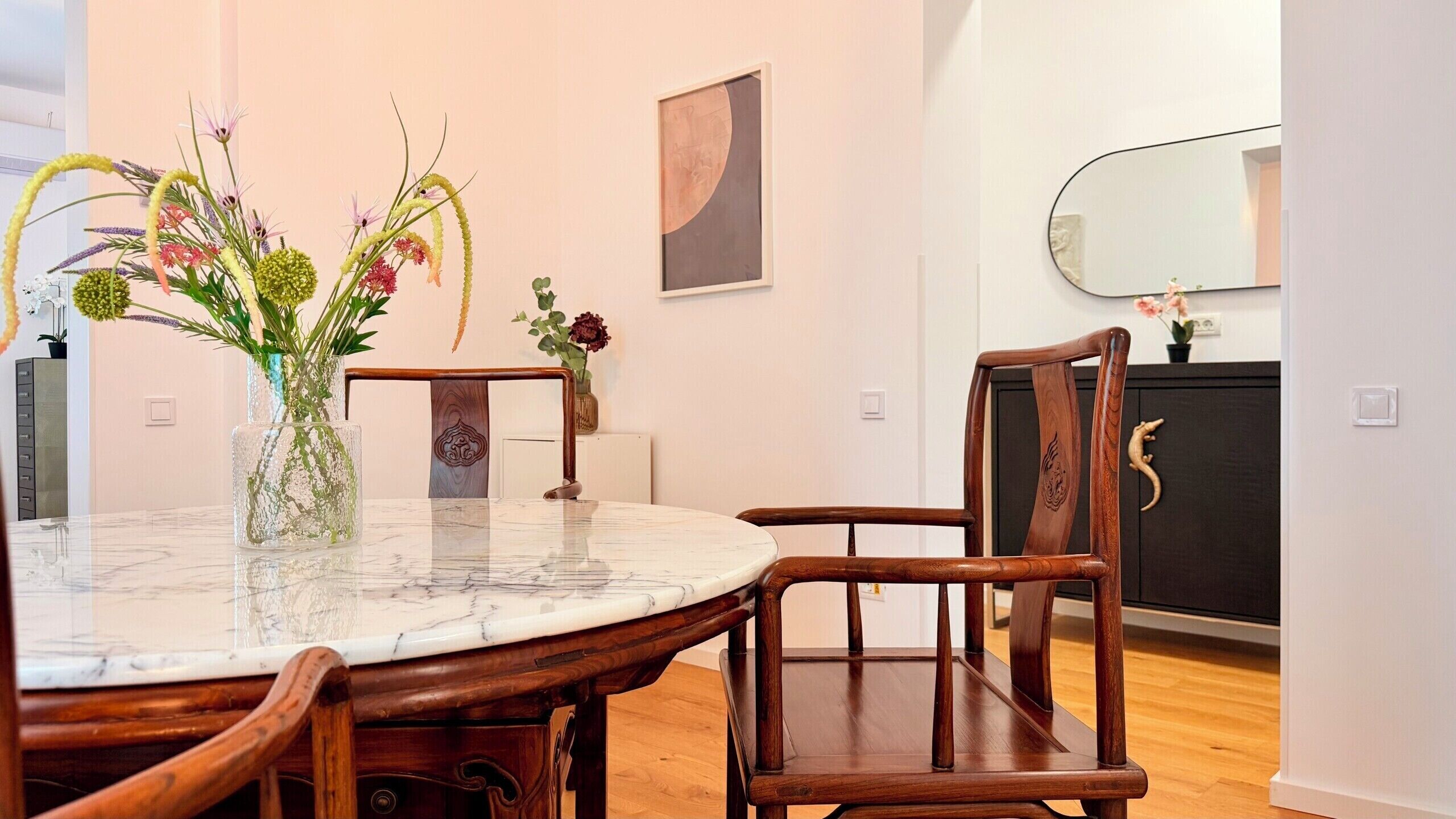Een eetgedeelte met een ronde marmeren tafel, houten stoelen en een vaas met bloemen.