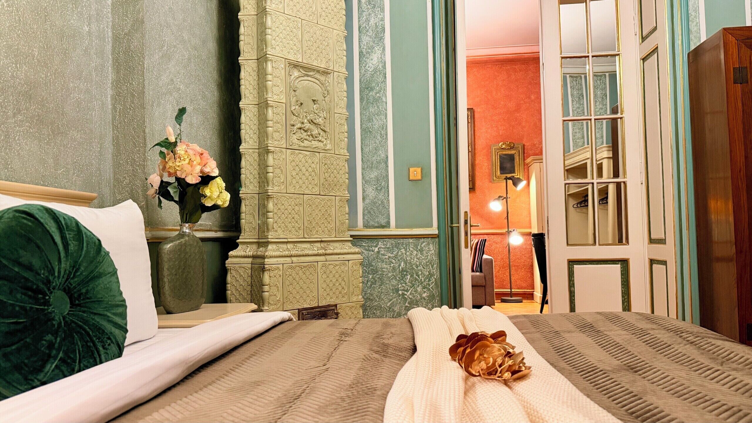 Habitación de hotel con cama, mesita de noche, un florero con flores, lámpara y vista al pasillo.