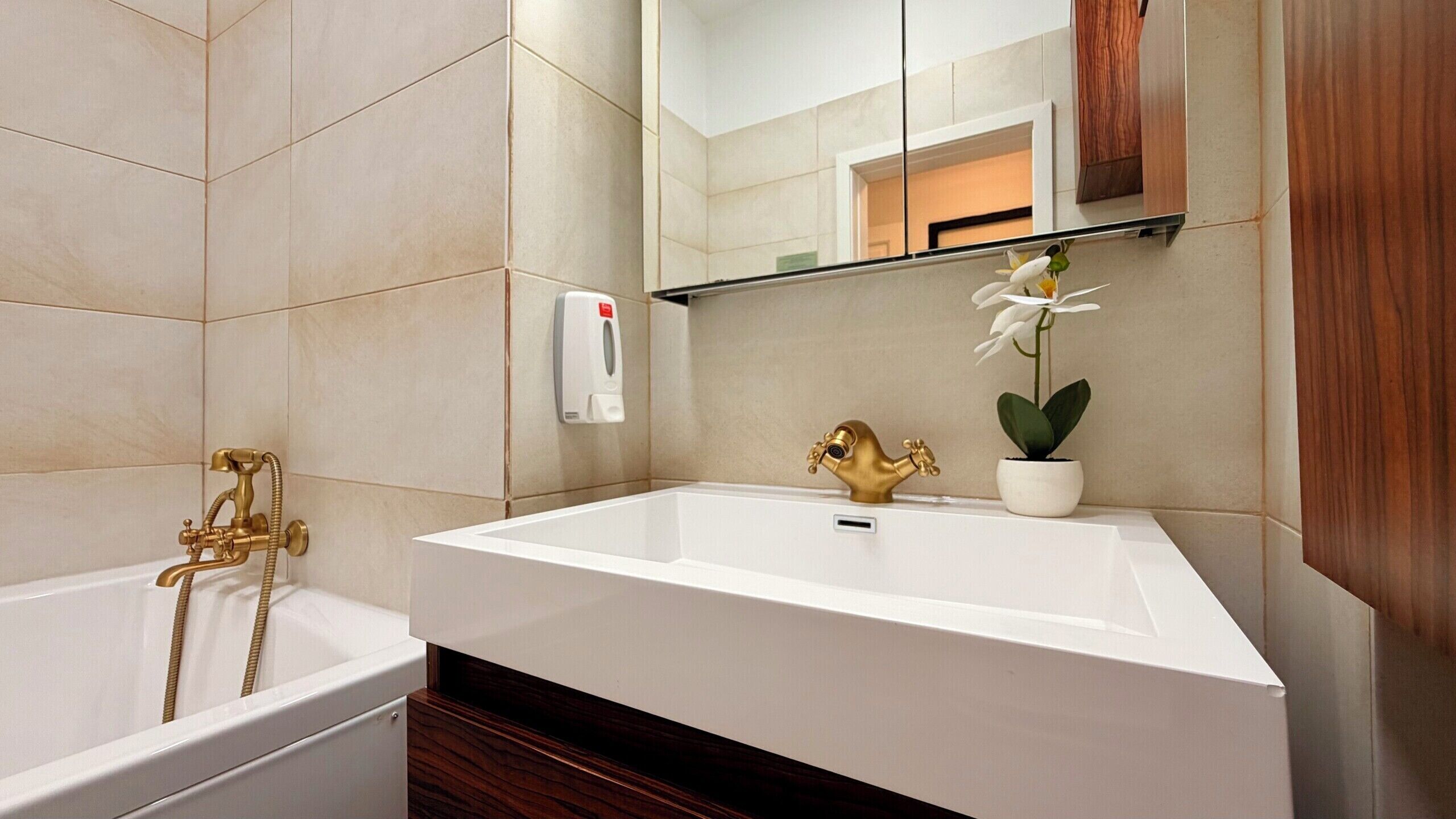 A modern bathroom with a large bathtub, a gold faucet, a white sink, and a potted orchid.