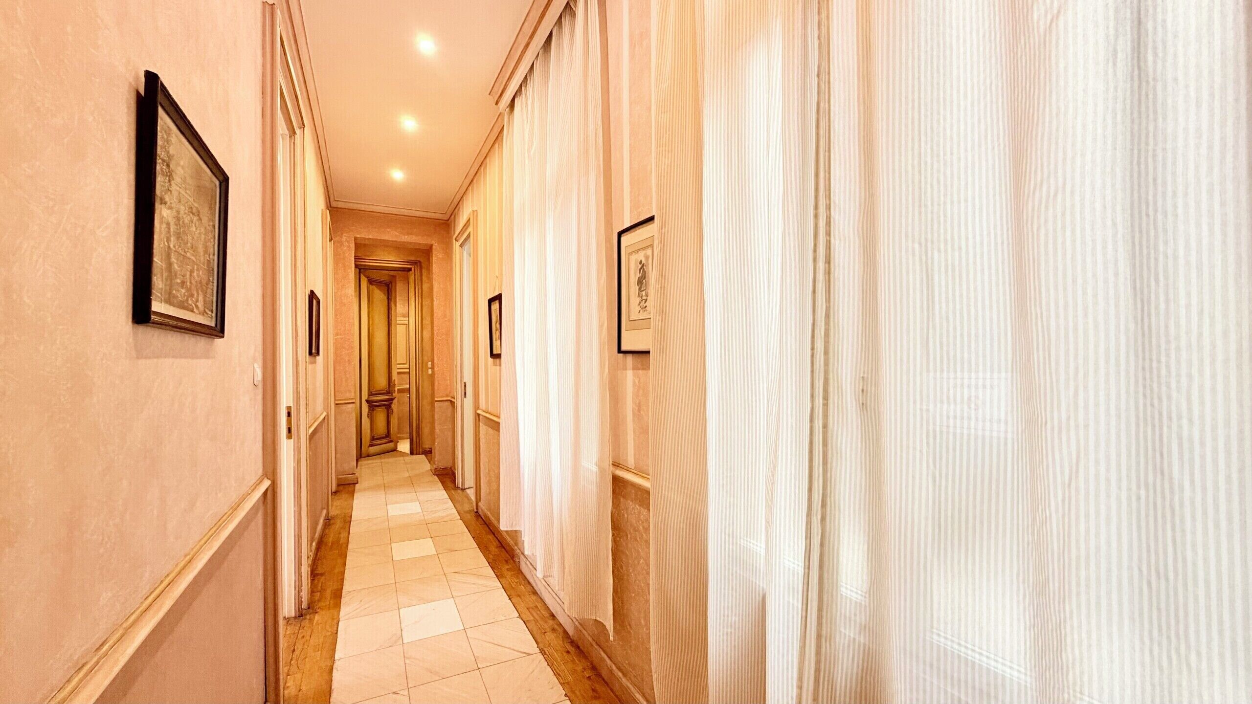 A hallway with framed pictures on the walls, a tiled floor, and a door at the end.