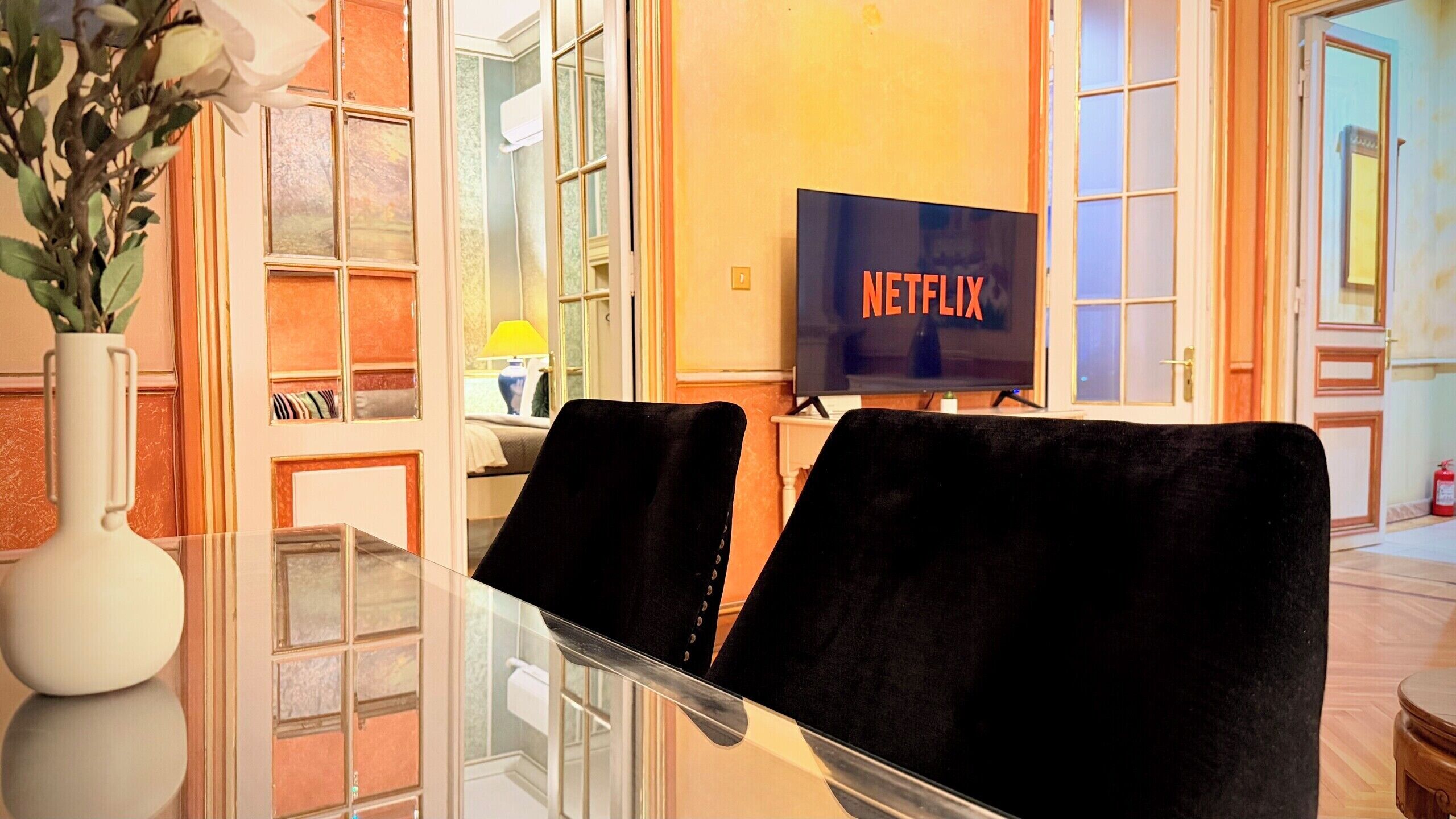 Una sala de estar moderna con una mesa de centro de vidrio, dos sillas de terciopelo negro, un jarrón blanco con flores y un televisor mostrando el logotipo de Netflix.