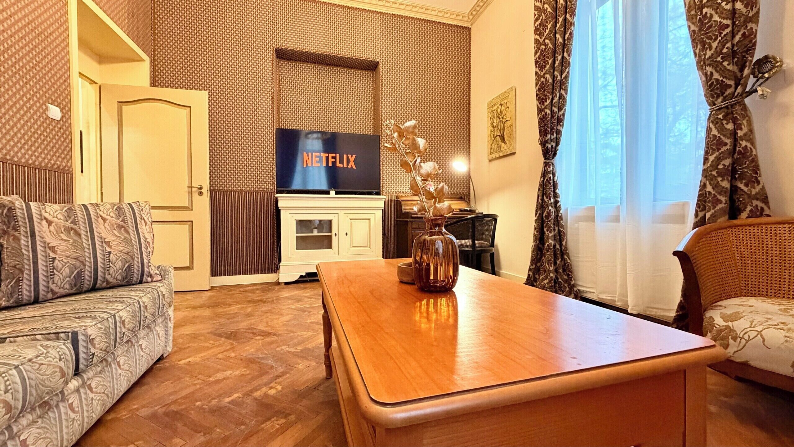 Una sala de estar con un sofá, una mesa de centro, un televisor mostrando 'NETFLIX' y una pared con diseño.