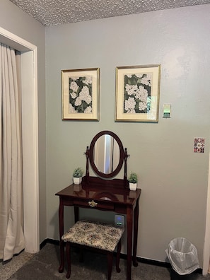 Een kamer met een kaptafel, twee ingelijste bloemwerk afbeeldingen, een spiegel, een kleine potplant en een prullenbak.