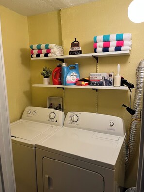 A laundry area with two front-loading washers, shelves with folded towels, a plant, and laundry supplies.