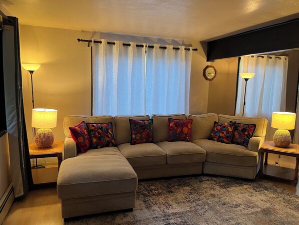 Una sala de estar con un sofá seccional color beige, dos mesas auxiliares con lámparas, un tapete estampado y un reloj de pared.