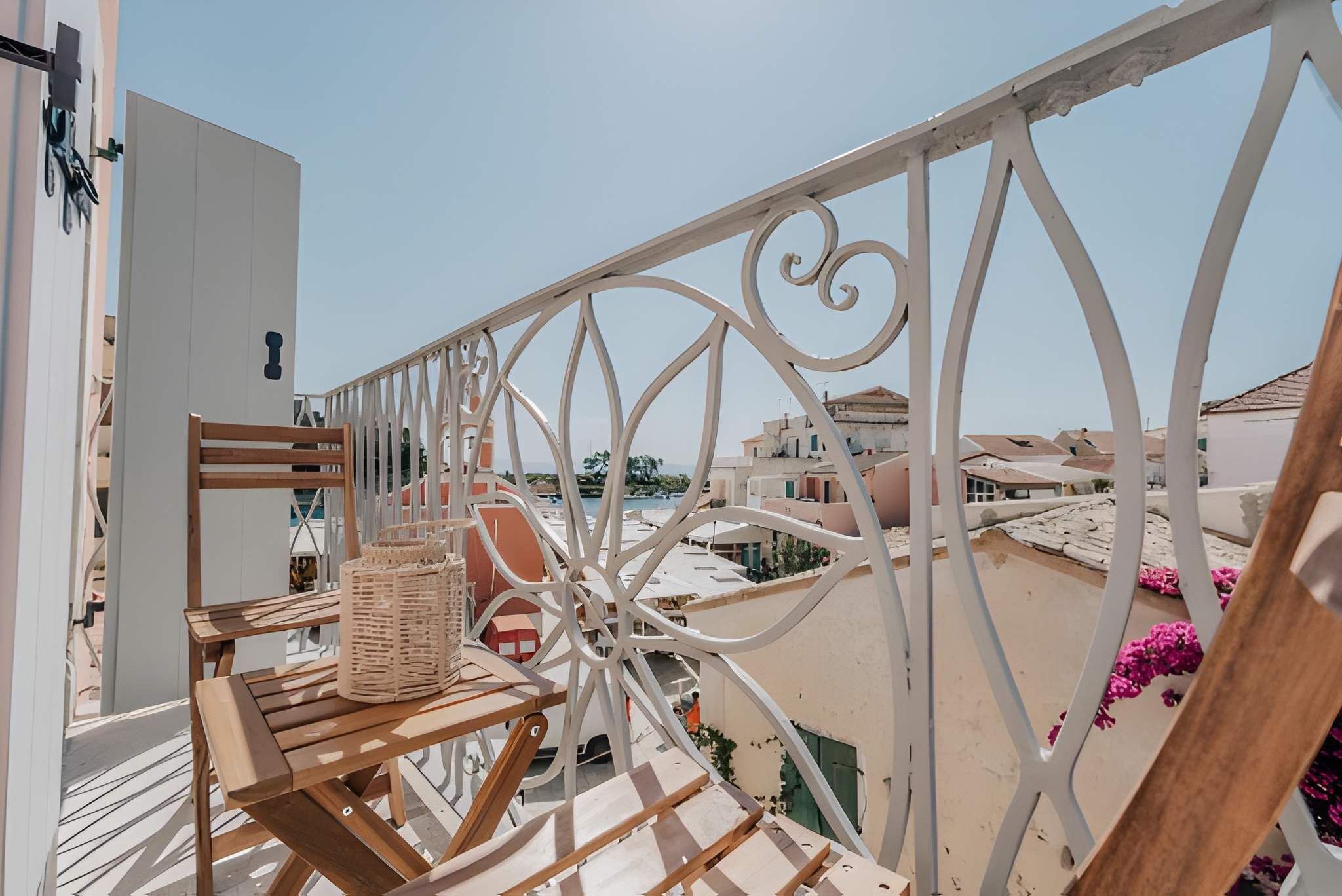 Un balcón con una silla de madera y una cesta de mimbre, con vistas a una zona residencial con edificios y cielo despejado.