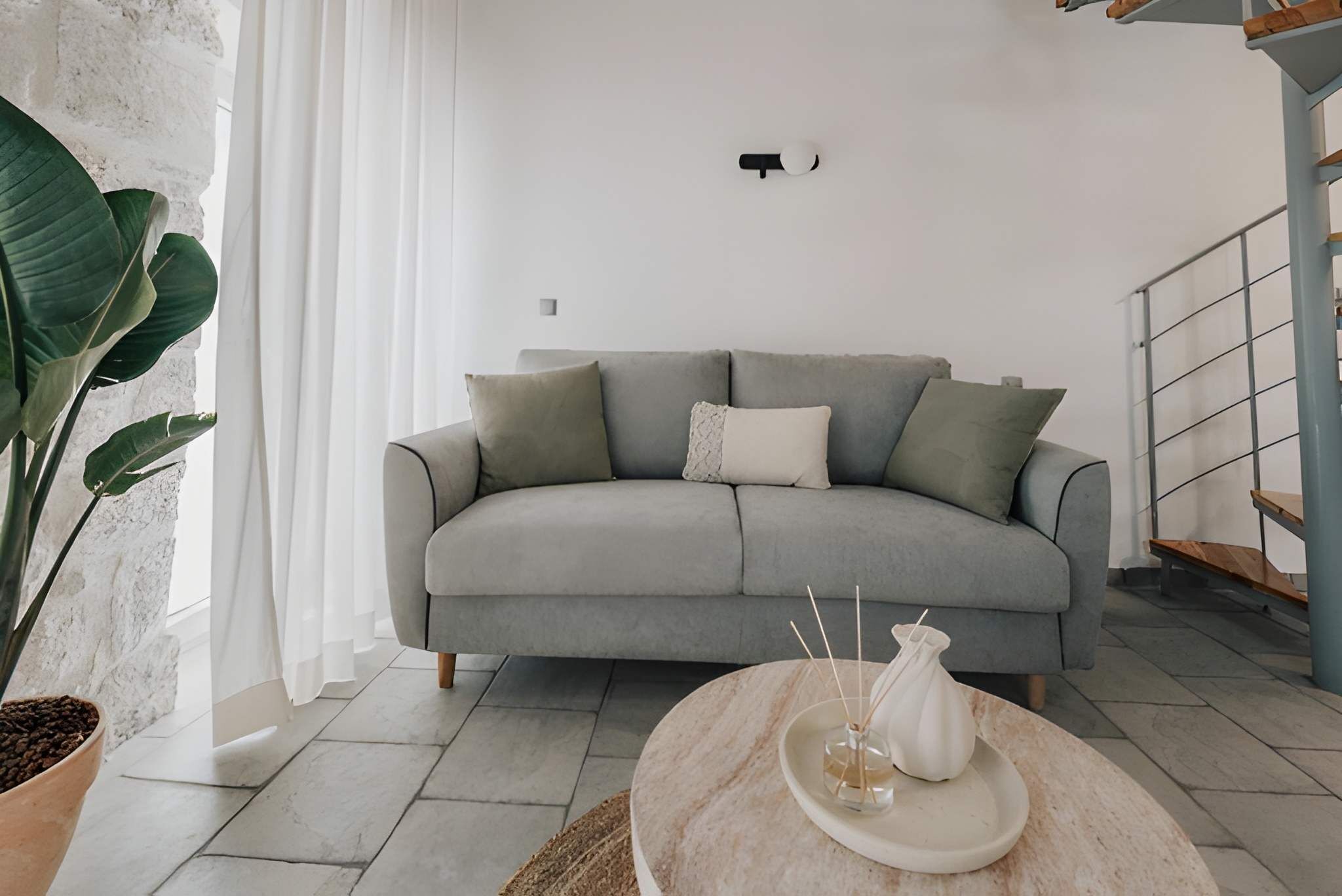 A modern living room with a grey sofa, a round table with a white vase, and a large plant.