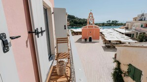 A balcony with a view of a coastal town, a building with a bell tower, and a paved area with umbrellas. - Luminis Residence by All about Paxos (Gaios)