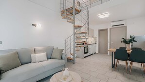 A modern living space with a light grey sofa, a round coffee table, a spiral staircase, a kitchen area, and a dining table with chairs. - Luminis Residence by All about Paxos (Gaios)