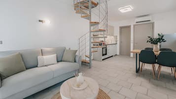 Un espacio habitable moderno con un sofá de color gris claro, una mesa redonda, una escalera de caracol, una zona de cocina y una mesa de comedor con sillas.
