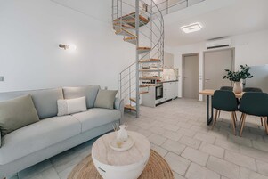 Un espacio habitable moderno con un sofá de color gris claro, una mesa redonda, una escalera de caracol, una zona de cocina y una mesa de comedor con sillas.