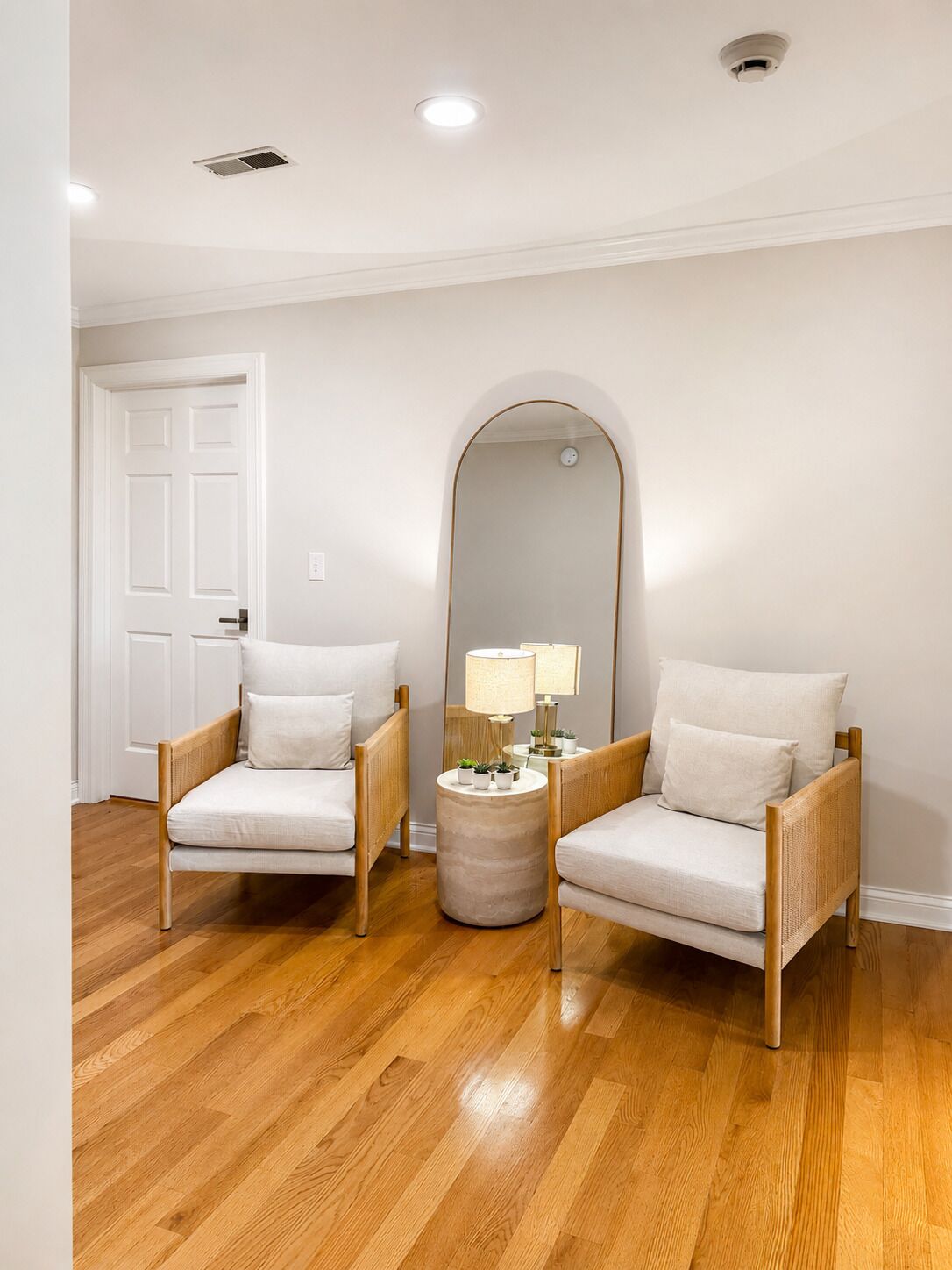 Une pièce avec deux chaises en bois, une petite table ronde avec une lampe, un grand miroir et une porte blanche.