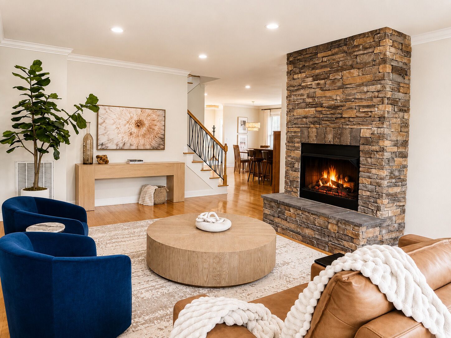 Una sala de estar con chimenea de piedra, sillones azules, una mesa redonda de madera y una escalera al fondo.