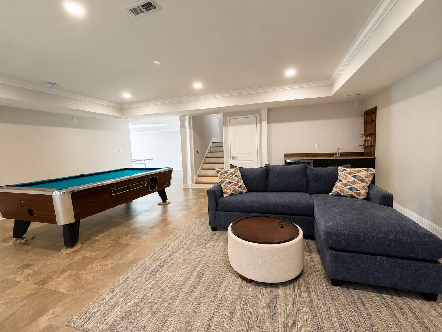Un salon moderne avec une table de billard, un canapé d’angle et un petit pouf rond.
