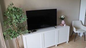 Un soggiorno con una TV a schermo piatto su un mobile bianco, una pianta in vaso, una sedia bianca e un piccolo vaso con fiori.
