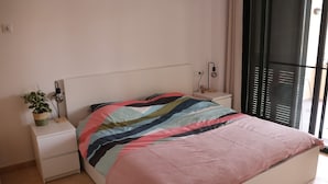 A bedroom with a bed, a bedside table with a lamp, a potted plant, and a window with blinds.