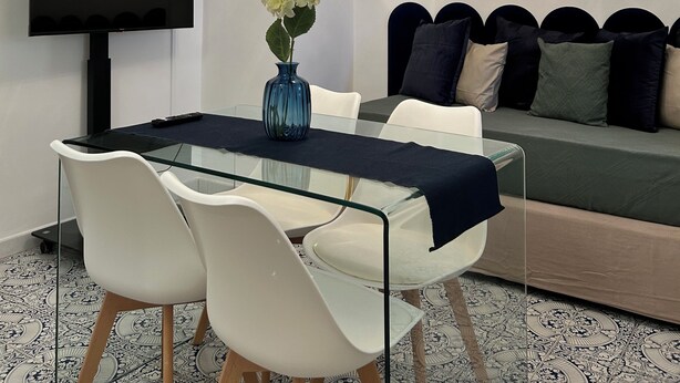 A modern living room with a glass dining table, white chairs, a sofa with dark cushions, a mounted TV, a decorative light fixture, and a framed artwork on the wall.