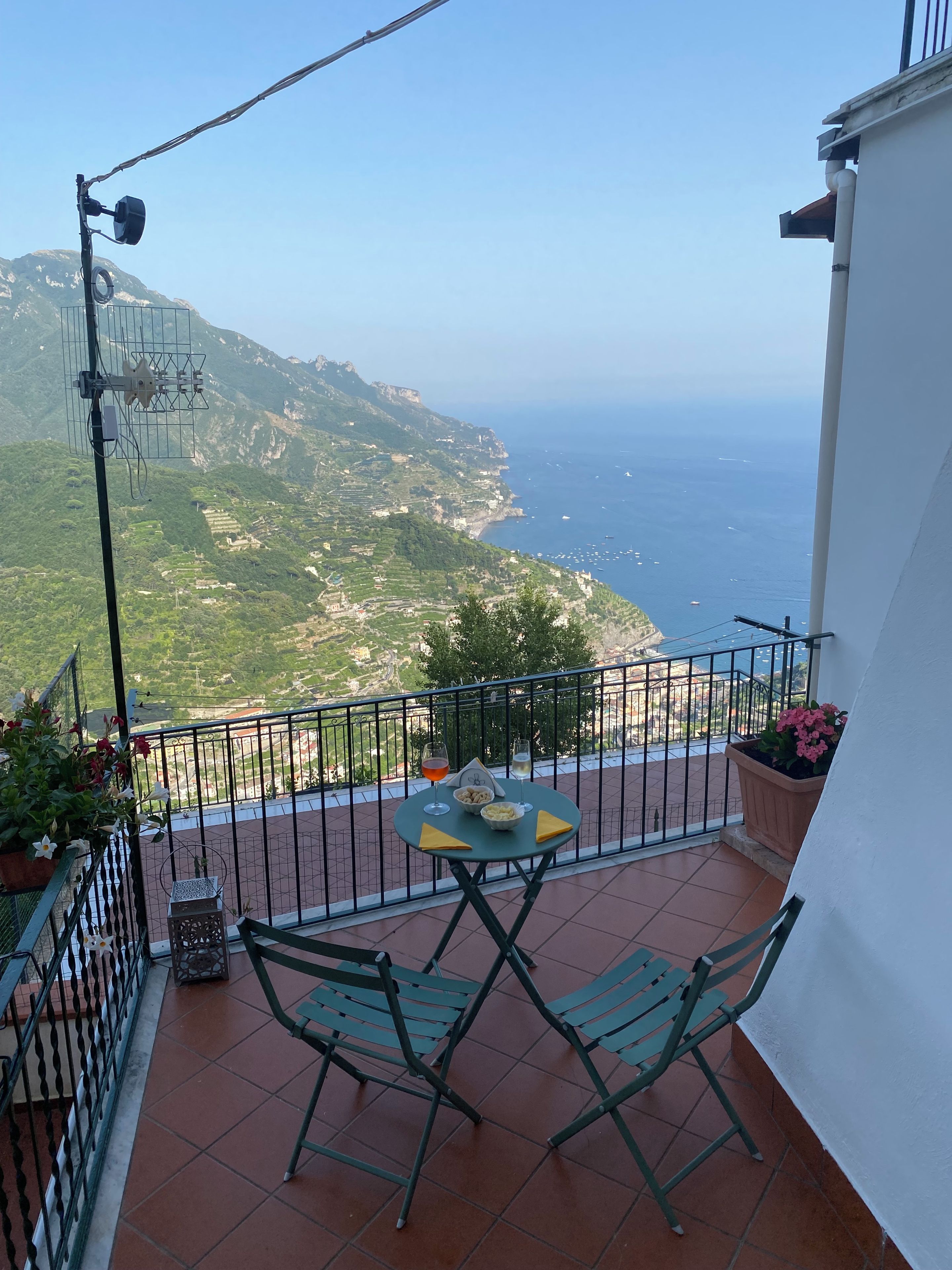 A balcony with a table and chairs, overlooking a coastal landscape with buildings and the sea.