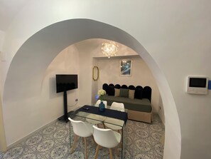 Una habitación con una mesa de vidrio, dos sillas blancas, un sofá con cojines, un televisor, un cuadro enmarcado y un termostato.