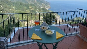 A balcony with a table set for two, overlooking a mountainous coastline with the sea in the distance.