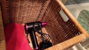 A wicker basket containing a red cloth and a black device with a pink handle, placed on a tiled floor near a window. - Casa Vitto 1-Room Apartment, A/C, 8 min from the airport, 10 min from the beach by car or bus (Genova)