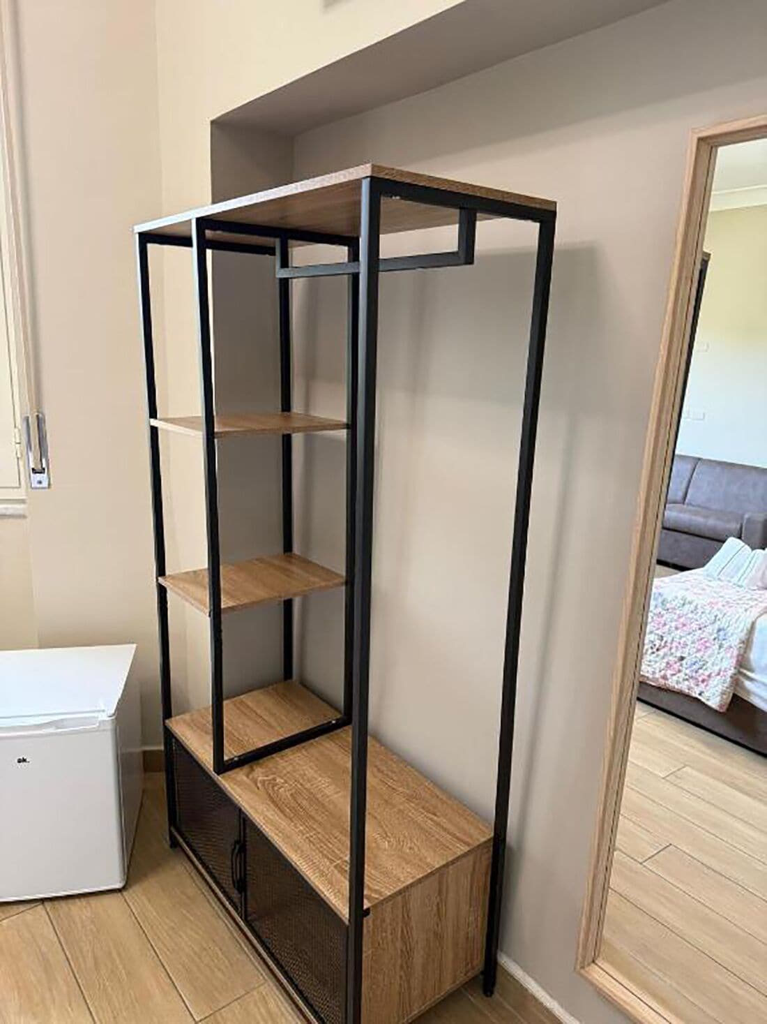 Une pièce avec une étagère haute et étroite le long du mur, un meuble blanc et un miroir qui reflète un lit recouvert d’une couverture à motifs.