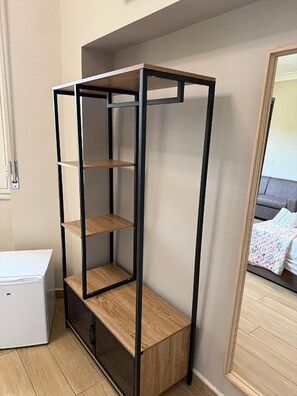 Una habitación con una estantería alta y estrecha contra la pared, un armario blanco y un espejo que refleja una cama con un edredón estampado.
