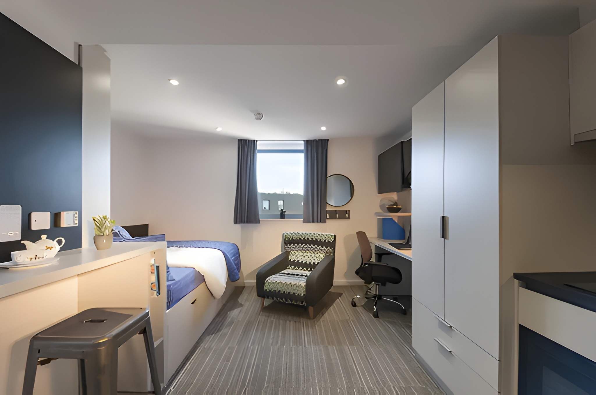 A compact hotel room with a bed, desk, chair, kitchenette, and a view of the city.