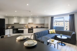 Eine moderne Küche mit eingebauten Geräten, ein Essbereich mit Tisch und Stühlen, ein graues Sofa und ein kleiner runder Tisch mit einer Pflanze.