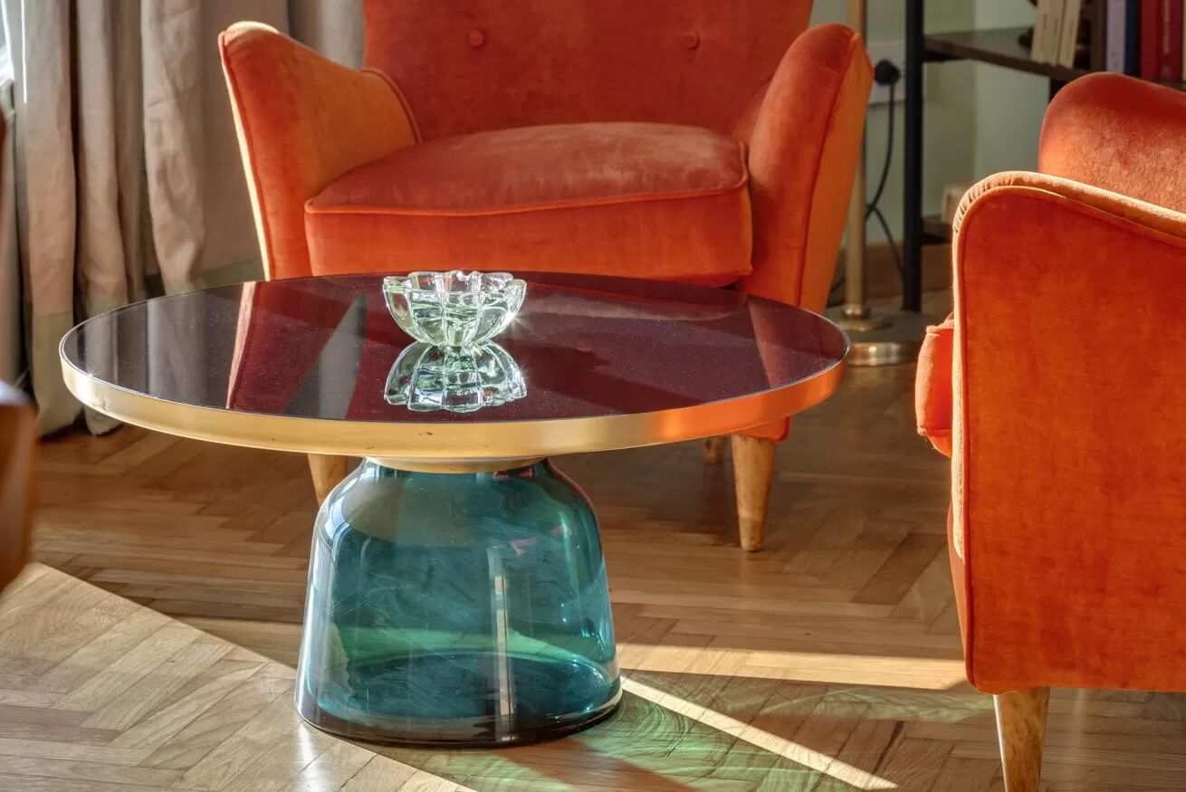 A round glass table with a metal base, a red velvet armchair, and a wooden floor.
