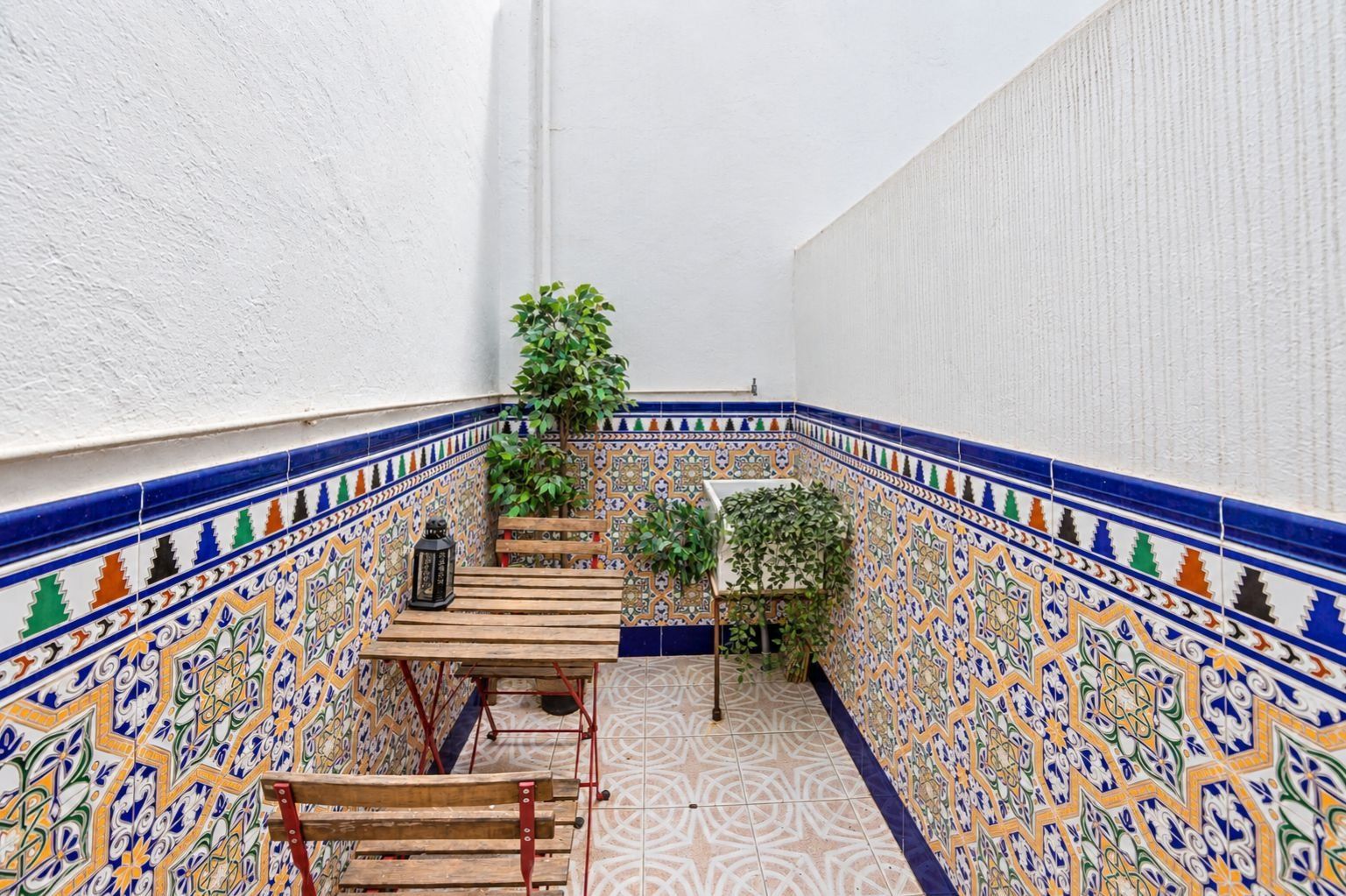A narrow outdoor corridor with a tiled floor, a wooden bench, a table, and a potted plant.