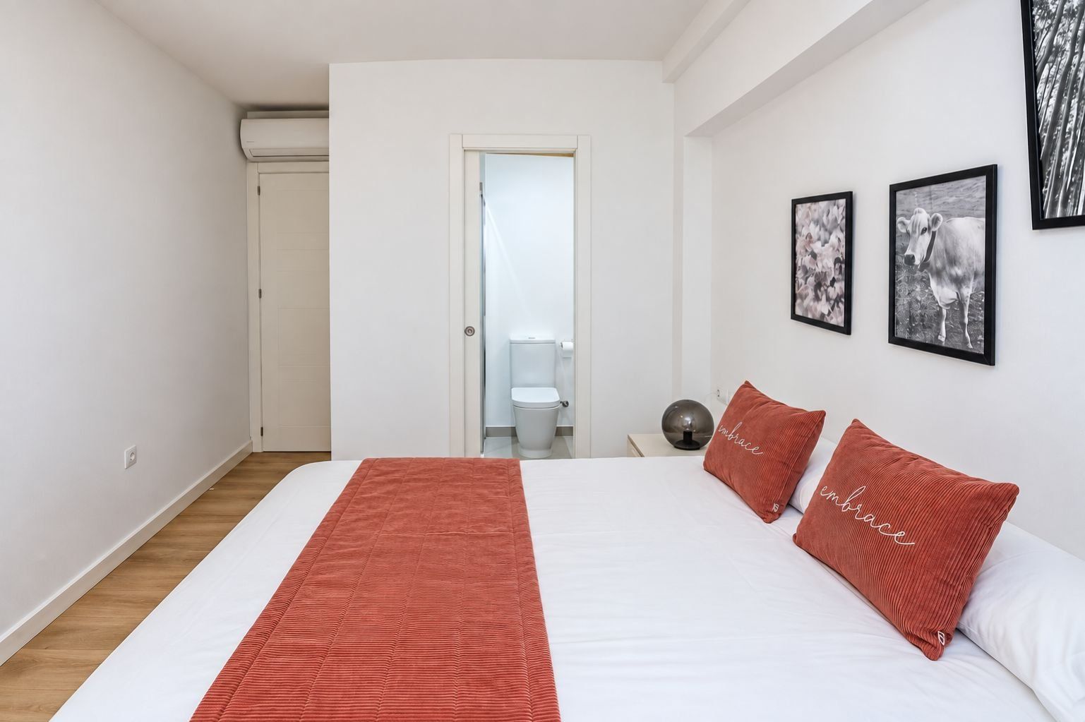A bedroom with a bed, a red throw blanket, two pillows with the word 'embrace,' a round table with a lamp, and a door leading to a bathroom.