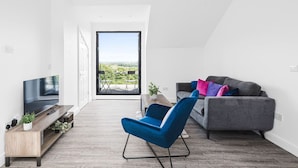 Ein modernes Wohnzimmer mit Fernseher, einem blauen Sessel, einem grauen Sofa mit bunten Kissen und einem Balkon mit Blick.