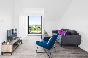 A modern living room with a TV, a blue chair, a grey sofa with colorful cushions, and a balcony with a view. - Panoramic View by MyCityHaven (Redland)