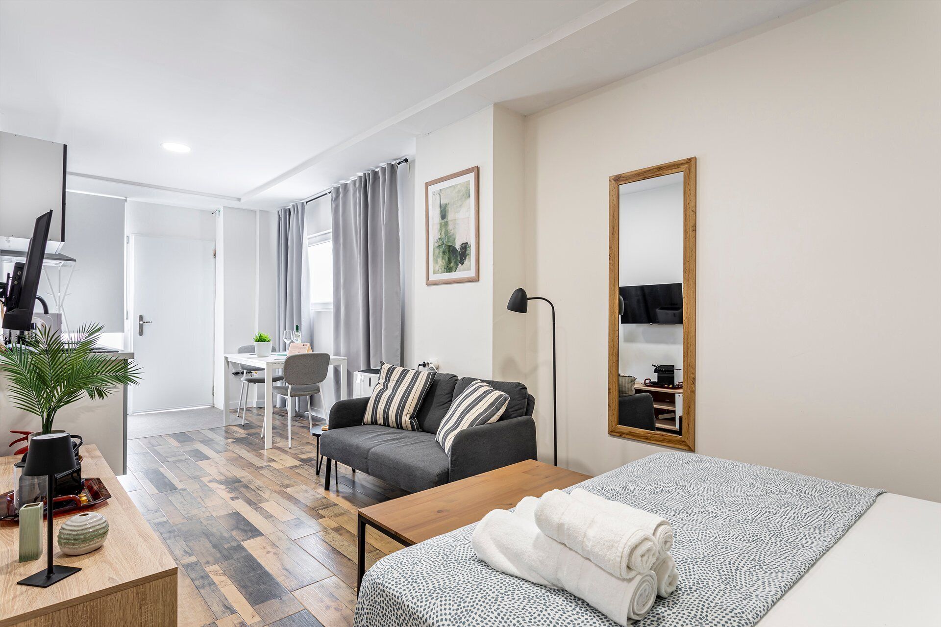 A modern hotel room with a bed, a desk, a chair, a sofa, a mirror, and a potted plant.