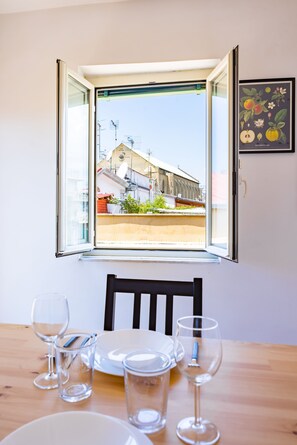 A dining table with chairs and wine glasses, a window with an open view of a building and greenery, and a framed botanical illustration on the wall. - Pazzariello Roof by NapoliApartments (Naples)