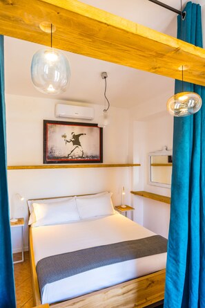 A bedroom with a wooden bed frame, a white mattress, a single bedside table with a lamp, a wall-mounted air conditioner, and a framed picture above the bed. - Pazzariello Roof by NapoliApartments (Naples)
