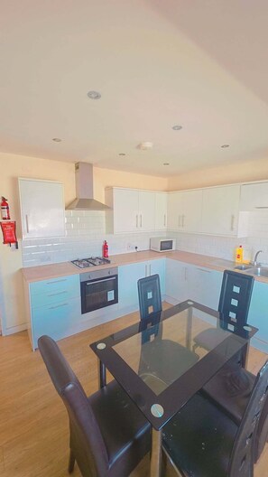 A modern kitchen with white cabinets, a gas stove, a microwave, and a glass dining table with chairs. - Heathrow ensuite rooms (Feltham)