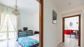 Un dormitori amb llit, una petita arca, televisió i finestra amb cortines.