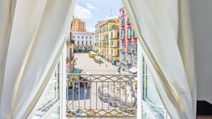 Vista des d'un balcó amb cortines blanques, sobre un carrer amb edificis de colors i una font.
