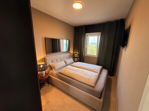 A bedroom with a bed, a nightstand, a lamp, a mirror, and a window with curtains. - Deluxe Sky Spa Penthouse & Resort (Toscolano Maderno)