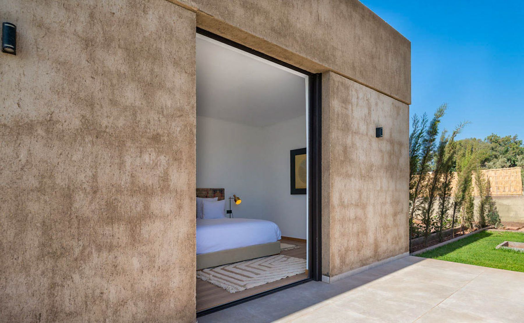 A modern bedroom with a large window, a bed, a nightstand, and a lamp.