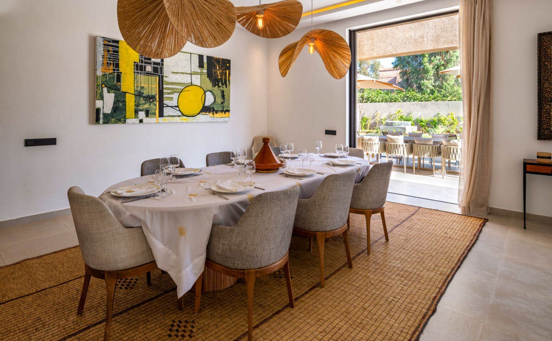 A dining area with a round table set for six, surrounded by chairs, with a large window offering a view of the outdoors.