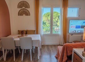 Ein Zimmer mit Esstisch, Stühlen, einem Fernseher und einem Fenster mit Blick auf Bäume.