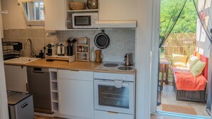 Eine kompakte Küche mit Mikrowelle, Herd und Spüle, ein kleines Holzregal mit Küchenutensilien und eine Glastür zu einem Außenbereich mit Sitzgelegenheit.