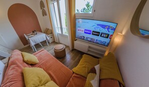 A living room with a television displaying various channels, a sofa with cushions, a small table with a bottle, and a window with curtains. - Charming studio with terrace near the beaches (Hyères)