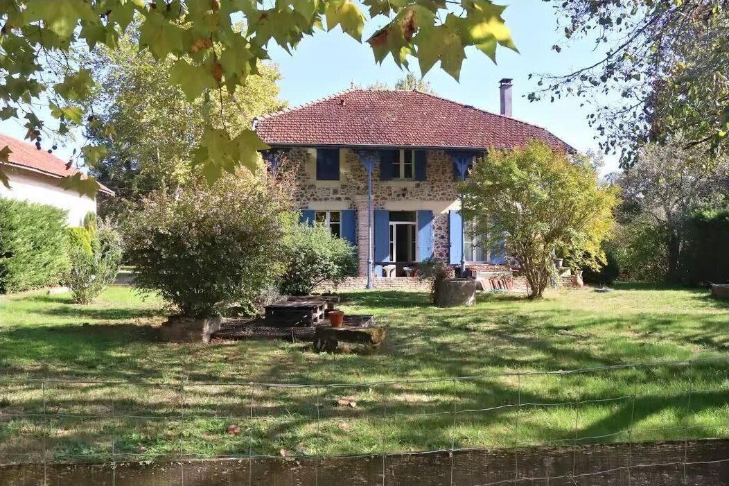 Une maison en pierre à deux étages, avec un toit recouvert de tuiles rouges, entourée d’une pelouse bien entretenue et d’arbres.