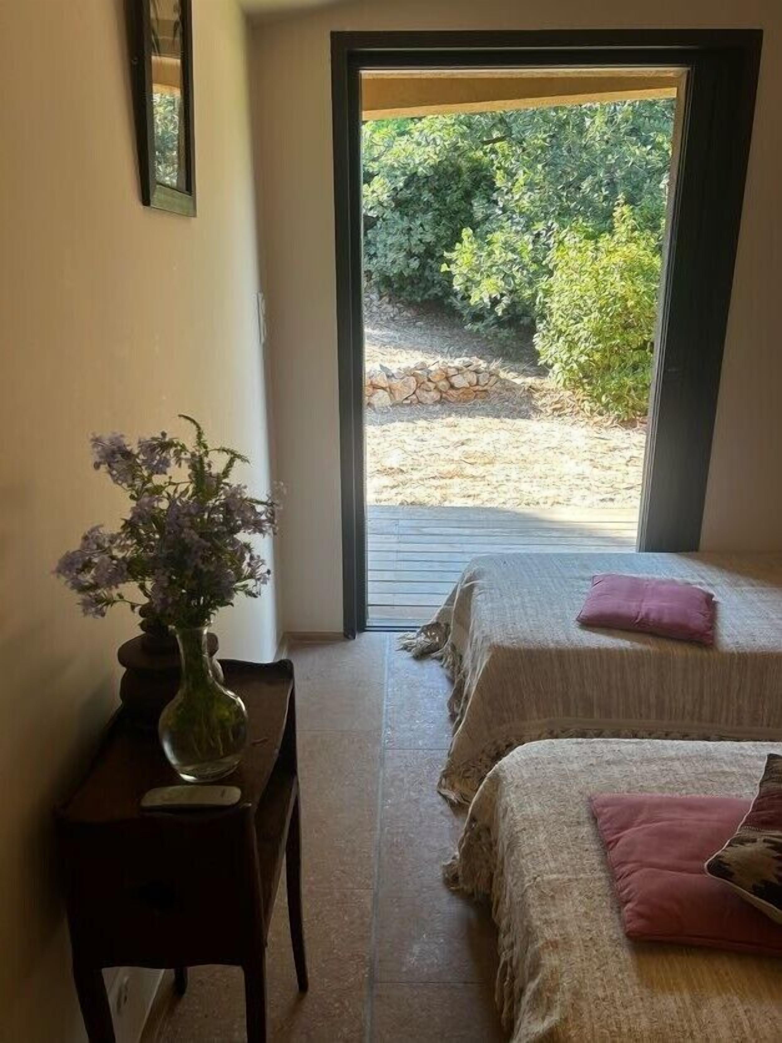 Un dormitorio con dos camas, una mesita de noche con un jarrón y vistas a un jardín exterior.